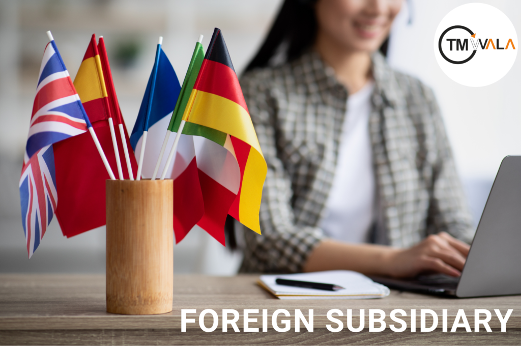 A desk with multiple international flags in a wooden holder, symbolizing foreign subsidiaries. A professional woman working on a laptop in the background, representing global business expansion